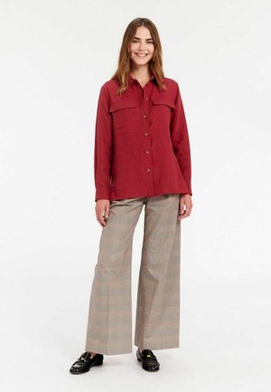 Chemise en lin rouge à boutons avec deux poches poitrine, associée à un pantalon large à carreaux gris et des mocassins noirs. Fond blanc.