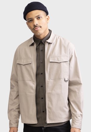 Young man wearing a beige jacket over a brown checkered shirt and a black beanie, standing against a light gray background.