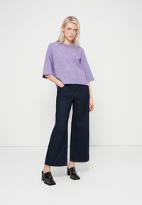 Pull violet à manches courtes associé à un jean large bleu foncé et des mules noires à talons carrés sur un fond neutre.