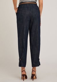 Los pantalones texturizados de color azul marino tienen dos bolsillos traseros, puños abotonados y un ajuste entallado, combinados con zapatos de tacón alto marrones.