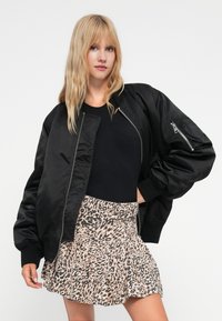 Veste bomber noire avec fermeture éclair sur le devant, poignets côtelés et texture lisse, associée à une jupe évasée beige à imprimé léopard.