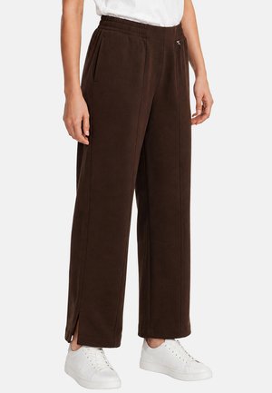 Persona che indossa pantaloni larghi marrone scuro con spacchi laterali e sneakers bianche, abbinati a una maglietta bianca, in piedi davanti a uno sfondo semplice.