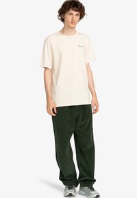 Beige cotton t-shirt with green text, paired with dark green corduroy pants. Model wears gray sneakers, standing against a plain background.
