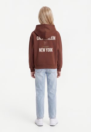 Personne aux longs cheveux blonds portant un sweat à capuche marron Calvin Klein New York, un jean bleu clair et des baskets blanches, de dos.