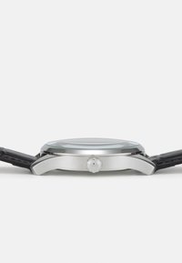 Reloj analógico con caja de acero inoxidable plateada, cristal curvado y correa de cuero negro. Presenta una corona prominente en el lateral.