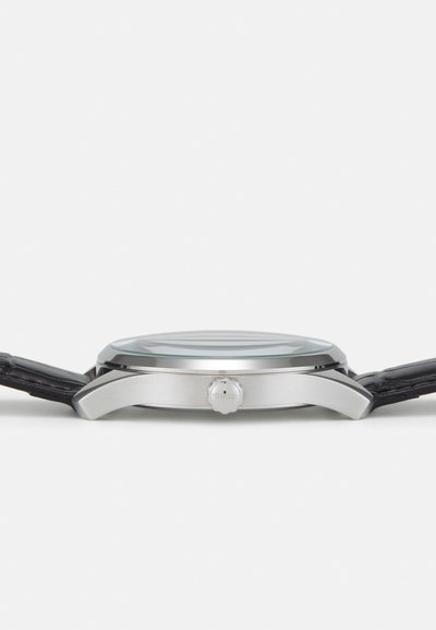 Montre analogique avec un boîtier en acier inoxydable argenté, verre courbé et bracelet en cuir noir. Elle dispose d'une couronne proéminente sur le côté.