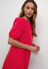 Ružičasta haljina kratkih rukava s napuhanim rukavima, okruglim izrezom i opuštenim krojem. Glatke teksture s minimalnim detaljima.