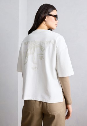 Γυναίκα που φορά γυαλιά ηλίου με σχέδιο καβουκιού, άσπρο φαρδύ T-shirt με μια μινιμαλιστική εικόνα προσώπου και το κείμενο «ΓΥΝΑΙΚΕΣ ΓΙΑ ΓΥΝΑΙΚΕΣ», και μπεζ παντελόνι.