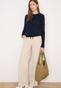 Granatowa bluza z kapturem ze ściągaczem u dołu, beżowe spodnie z szerokimi nogawkami oraz oliwkowa torba. Stylizację dopełniają białe trampki z czerwonymi akcentami.