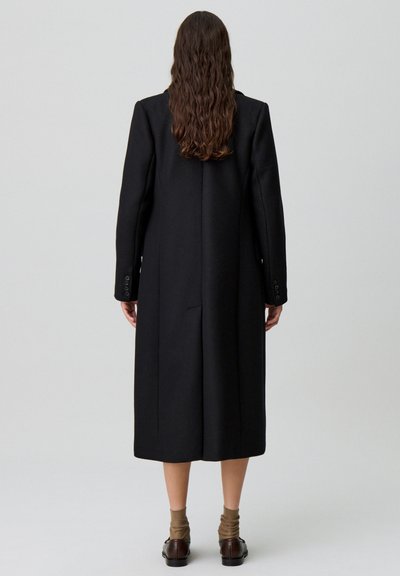 Cappotto lungo in lana nera con taglio dritto, spacco singolo sul retro e maniche con bottoni. Il design è minimalista e su misura.