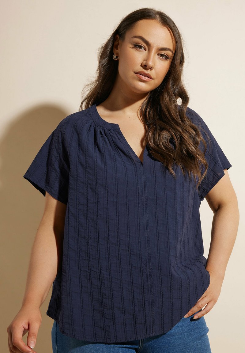 Blusa corta blu navy a maniche corte con scollo a V e linee di texture verticali, caratterizzata da una vestibilità rilassata e leggeri plissee alla scollatura.