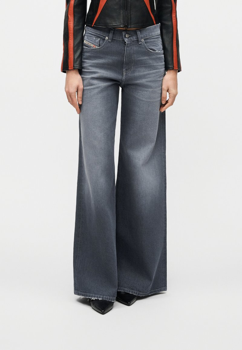 Γυναίκα που φοράει φθαρμένα γκρι τζιν παντελόνια με φαρδιά μπατζάκια, μαύρα μυτερά παπούτσια και μαύρο δερμάτινο μπουφάν με πορτοκαλί ρίγες στα μανίκια.