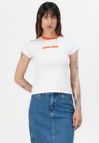 White cotton t-shirt featuring an orange ribbed collar and logo print. Short sleeves and fitted design. Paired with a denim skirt.