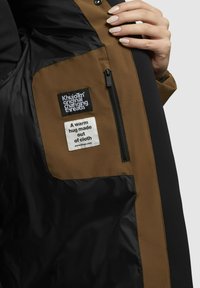 A barna és fekete dzseki belsejében zipzáros zseb, szőtt és nyomtatott címkék találhatók szöveggel, valamint sima, fényes textúrák jellemzik.