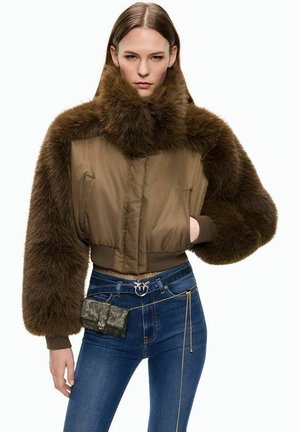 Donna che indossa una giacca corta in pelliccia marrone con maniche oversize e jeans blu attillati, tenendo una piccola borsa a catena in vita.
