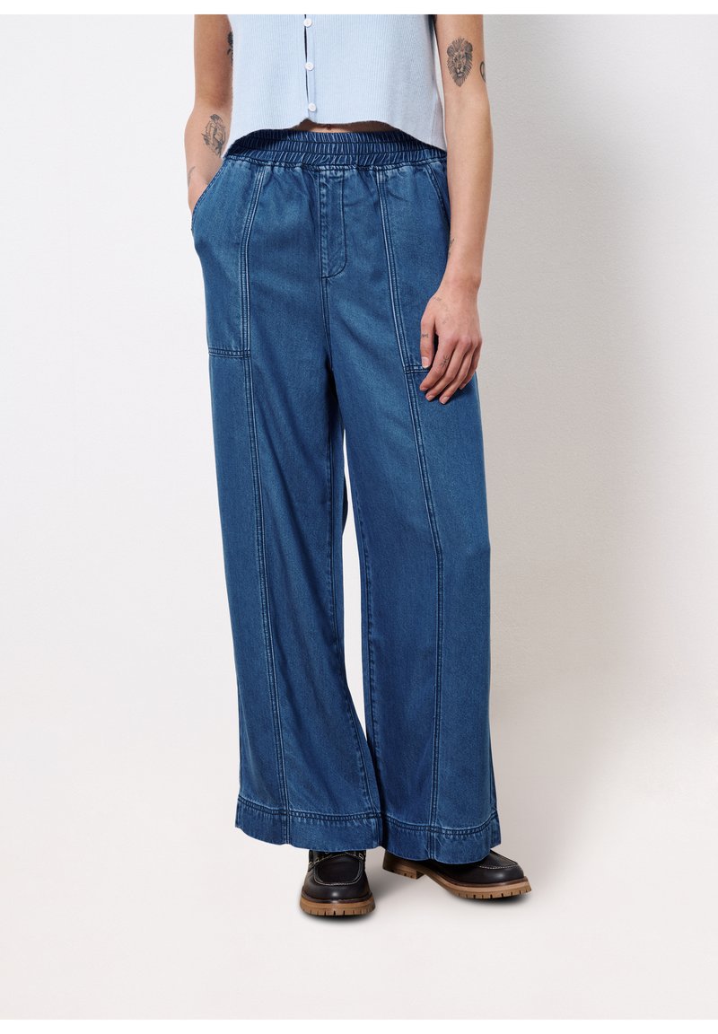 Pantalons large en denim bleu avec une taille élastique, des coutures droites et des poches latérales, associés à des chaussures sombres. Texture lisse et coupe ample.