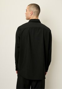 Chemise noire à manches longues avec col, en tissu texturé. Le dos présente un ourlet arrondi et des poignets boutonnés pour un ajustement réglable.