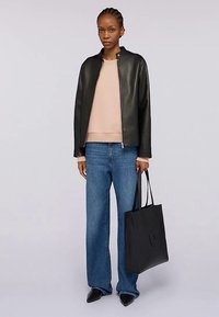 Chaqueta de cuero negra, sudadera rosa claro, vaqueros de pierna ancha azules, bolso tote negro con logo y zapatos negros puntiagudos. Modelo posando frente a un fondo neutro.