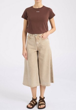 Femme portant un t-shirt marron à manches courtes avec le texte « PINKO », un pantalon beige large et court, et des sandales marron à brides, debout avec une main dans la poche.