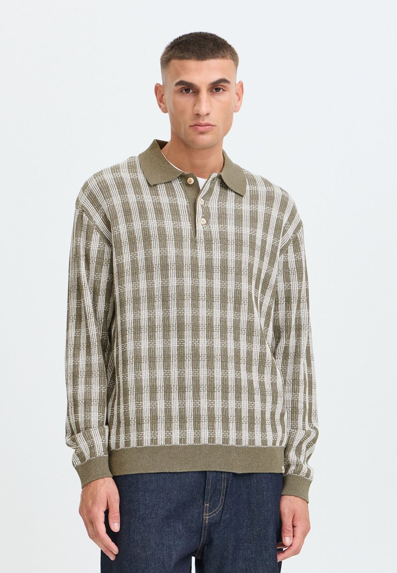 Homme portant un polo en maille à manches longues à carreaux verts et blancs avec des boutons et un jean bleu foncé, debout devant un fond uni.