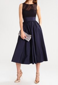 Navy ärmelloses Kleid mit einem transparenten schwarzen Oberteil, einem plissierten Rock und einer taillierten Taille. Kombiniert mit einer silbernen Clutch und goldenen Riemchen-Heels.