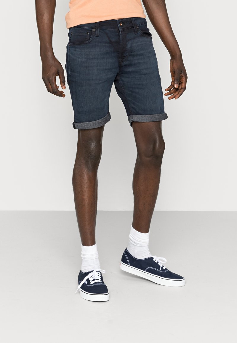 Dark denim shorts with rolled cuffs, featuring classic five-pocket design. Worn with white socks and navy sneakers. Smooth texture.