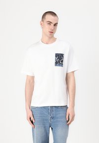 Camiseta de algodón blanca con mangas cortas y cuello redondo; presenta un bolsillo con un patrón azul y negro en el lado izquierdo del pecho.