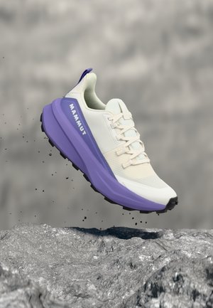 Chaussure de trail running Mammut blanche et violette en plein air au-dessus d'un terrain rocheux, avec une semelle noire et des lacets blancs.