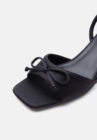 Μαύρο πέδιλο με ανοιχτά δάχτυλα, τετράγωνη μπροστινή όψη, χαμηλό τακούνι και διακοσμητικό φιόγκο δεμένο στο λουράκι που περνάει πάνω από τα δάχτυλα.