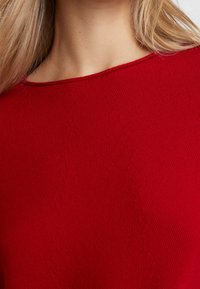 Pull en tricot rouge avec un col large, un tissu texturé et des détails de coutures subtils. Texture lisse sans ornements.