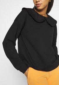 Felpa nera con colletto arricciato, maniche lunghe, vestibilità comoda e polsini a costine, abbinata a pantaloni arancioni.