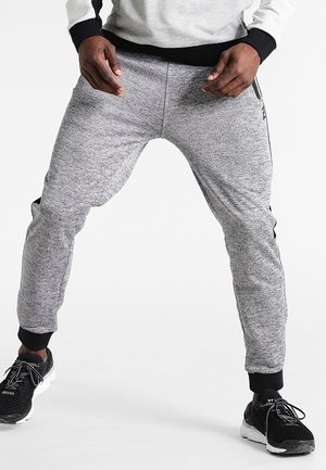 Man in grijze joggingbroek en zwarte sneakers met gespreide benen op een witte achtergrond.