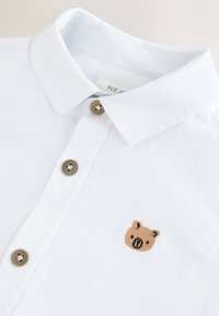 Polo blanc avec des boutons marron et un petit visage d’ours marron brodé sur la poitrine près des boutons.