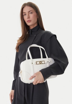 Donna con abito nero a maniche lunghe che tiene una borsa bianca con dettagli dorati e logo A|X su uno sfondo bianco uniforme.