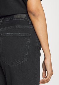 Melna denim džinsi ir ar klasisko piecu kabatu dizainu, redzamu šuvju un plāksteri uz jostasvietas, ar gludu tekstūru.