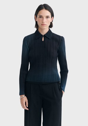Femme aux cheveux courts et foncés portant un haut côtelé bleu foncé à manches longues avec un col et un pantalon noir sur un fond clair uni.