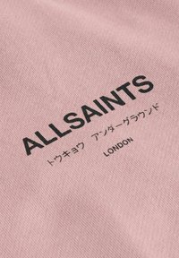 Svetlo ružová tkanina s čiernym textom "ALLSAINTS" a ďalšími japonskými znakmi, s hladkou textúrou a jednoduchým dizajnom.