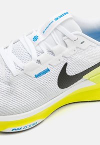 Baskets de course blanches avec un dessus en mesh texturé, logo Nike noir, accents bleus et une semelle jaune vif avec l'inscription "Zoom".