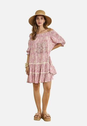 Mujer con vestido rosa con flores y hombros descubiertos con volantes, un sombrero de paja de ala ancha, pulseras doradas y sandalias de plataforma, posando frente a un fondo blanco.