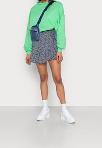 Grüner oversized Hoodie, marineblauer Minirock mit Rüschendetails, weiße Plateausneaker und eine kleine blaue Umhängetasche mit einem Trageriemen.