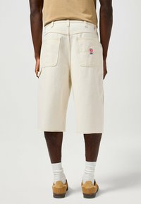 Pantalones cortos de carga beige con dos bolsillos traseros, costuras en contraste y un pequeño parche de marca. El material parece duradero y ligeramente texturizado.