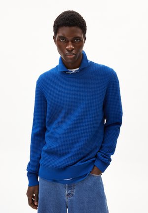 Pull en maille bleu texturé avec un col haut et des poignets côtelés, porté sur une chemise blanche, associé à un short en denim bleu clair.