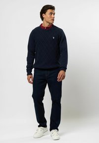 Junger Mann steht, trägt einen marineblauen Pullover mit Rautenmuster über einem roten Hemd mit Kragen, dunkle Jeans und weiße Sneakers, schaut nach rechts.