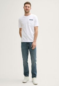 Hvid T-shirt med korte ærmer og brystlomme, parret med blå denimjeans. Modellen har hvide sneakers på, med en ensfarvet baggrund.