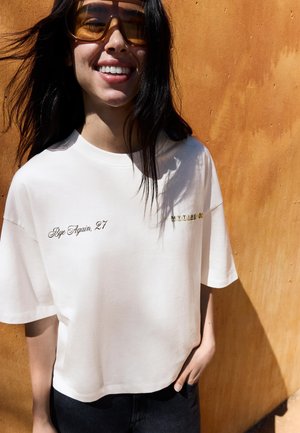 Lächelnde Frau mit langen dunklen Haaren trägt übergroße bernsteinfarbene Sonnenbrille und ein weißes T-Shirt mit schwarzen Hosen, steht vor einer orangenen Wand.