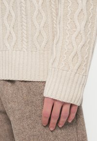 Bézs színű kötött pulóver bordázott mandzsettákkal, dombornyomott mintákkal és puha, vastag megjelenéssel, barna texturált nadrággal párosítva.