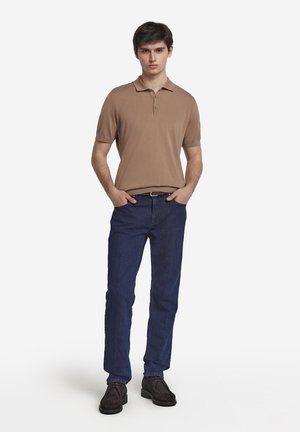 Brązowa koszulka polo, ciemnoniebieskie dżinsy i ciemne buty. Koszulka ma kołnierzyk i krótkie rękawy. Dżinsy mają klasyczny krój.