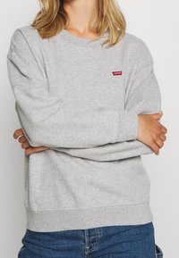 Szara bluza z miękką fakturą, ściągaczami na mankietach i dole oraz małym czerwonym logo Levi's na piersi. Casualowy design.