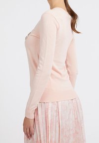 Jasnoróżowy, długi sweter z miękkiego materiału, z prążkowanym dołem i mankietami. Na wysokości klatki piersiowej znajduje się ozdobny element.