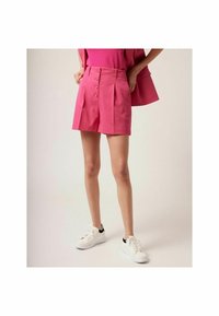 Shorts taille haute roses avec fermeture à boutons sur le devant, plis et finition texturée. Associés à des baskets blanches avec des accents noirs.